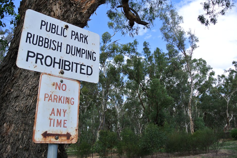 Close-up view of two weathered metal signs affixed to a rough tree trunk in a natural park setting. The upper sign is white with bold black text reading 'PUBLIC PARK RUBBISH DUMPING PROHIBITED,' indic